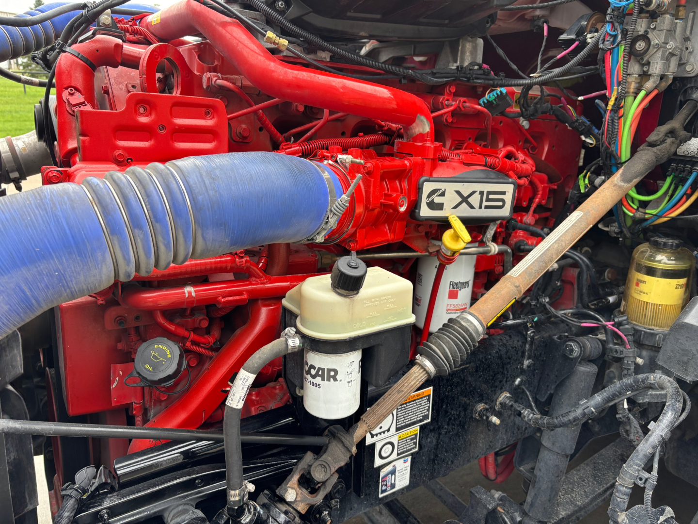 Kenworth engine bay showing X15 emblem, blue intercooler hose, white reservoir, and red engine components in a sleeper tractor