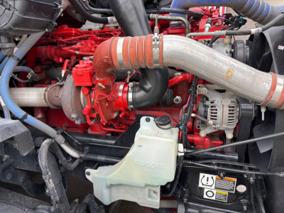 Kenworth T680 engine bay showing red engine block, large silver intake pipe, coolant reservoir, and alternator with wiring.