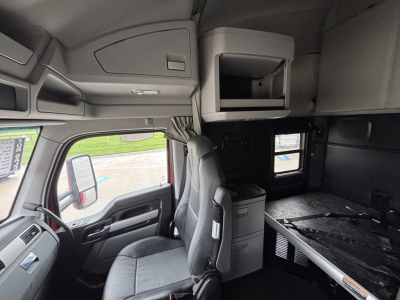 Kenworth T680 truck interior cabin view showing sleeper area, cabinetry, and small fridge area. Front passenger seat visible with steering wheel nearby, side mirror visible, window and dashboard components.