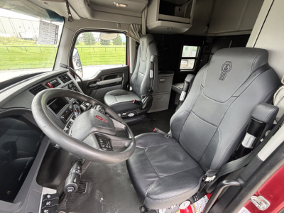 Kenworth truck interior driver area with steering wheel, large grey seats, dash controls; appears to be a modern cab with overhead storage and window to the left.