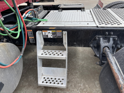 Two-step aluminum foothold attached to a Kenworth T680 truck chassis with perforated steps and warning labels, wiring harnesses, and a portion of the drive axle visible.