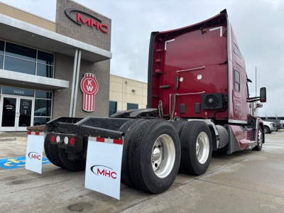 Kenworth T680 truck chassis with red sleeper cab, rear view shows dual rear axles and tires, MHC dealership building in background.