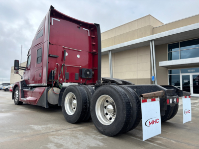 Kenworth T680 semi truck rear/side view showing rear axles and mud flaps with MHC sign, maroon cab and fifth wheel, concrete lot and building in background.