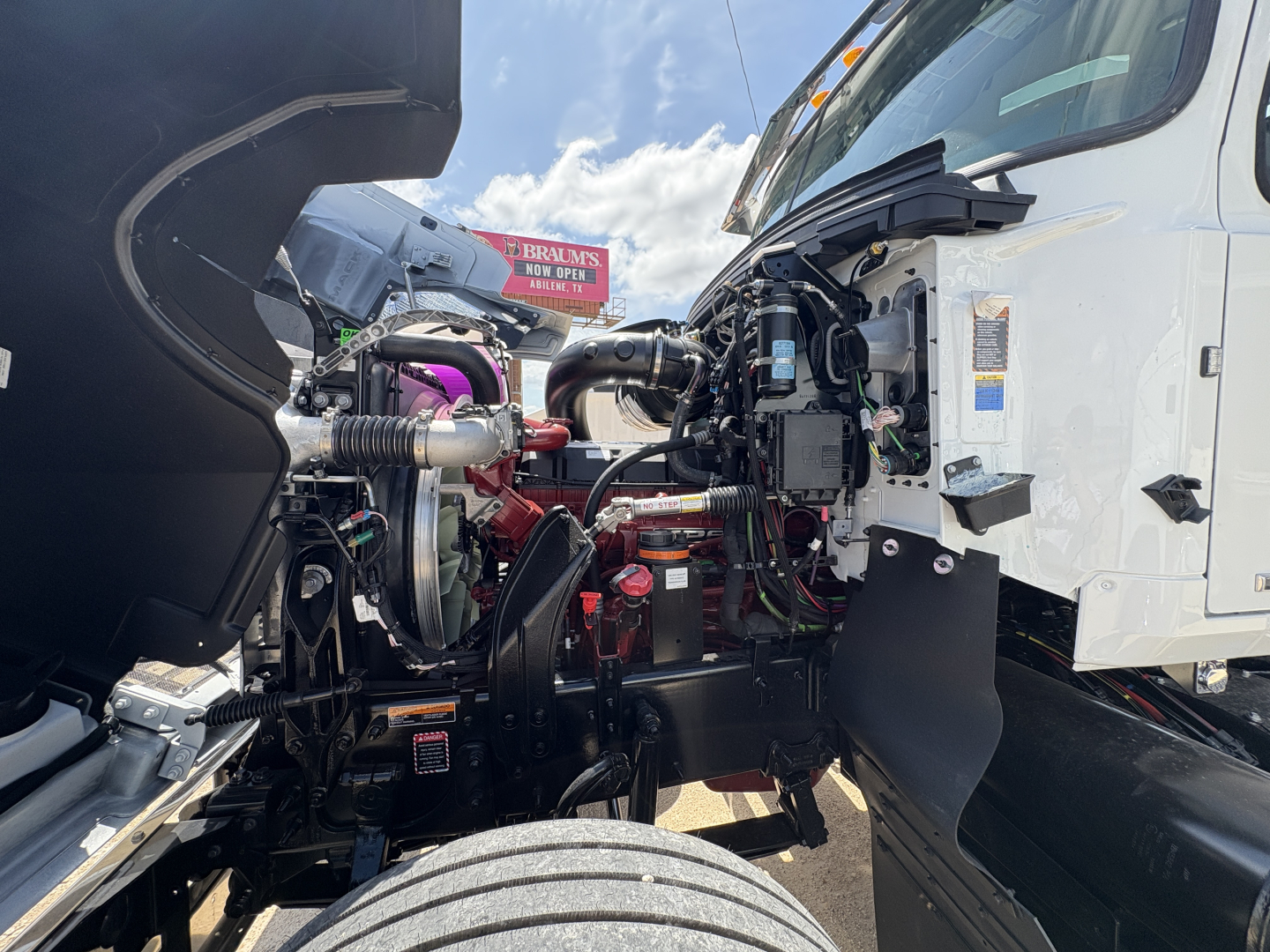 Front engine bay of a 2027 Mack PI64T truck with open hood; visible components include turbocharger, hoses, wiring, intake, and red engine parts; cab interior not visible.