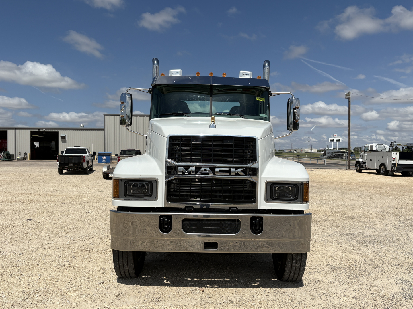 Mack truck front view with large grille, 'MACK' emblem, white cab, chrome bumper, and dual exhaust stacks. Clear sky with a few clouds.