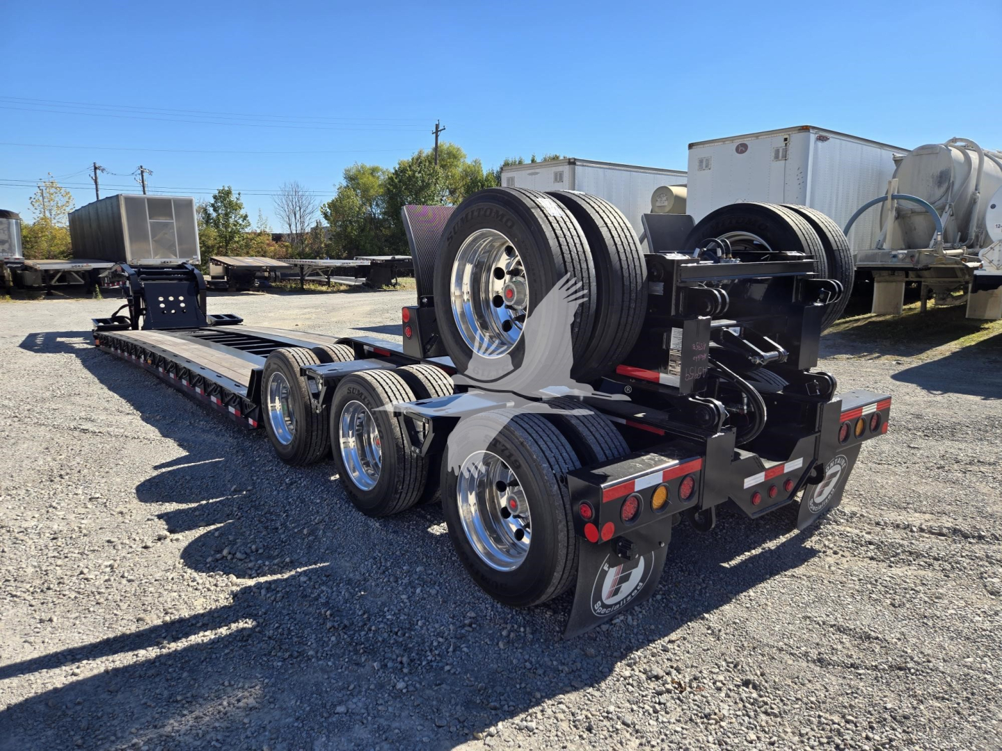 Semi-trailer with extendable lowboy style deck showing multiple axles and a mounted spare tire; no visible text on signs; background includes other trailers and equipment.