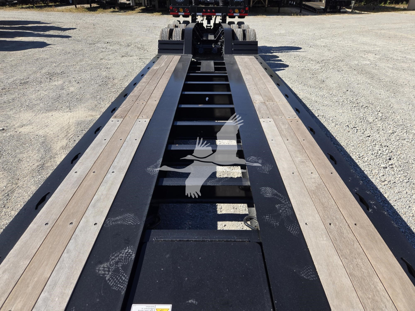Lowboy trailer deck with long steel frame and wooden planks, hydraulic flip mechanism visible at far end, no cab in view.