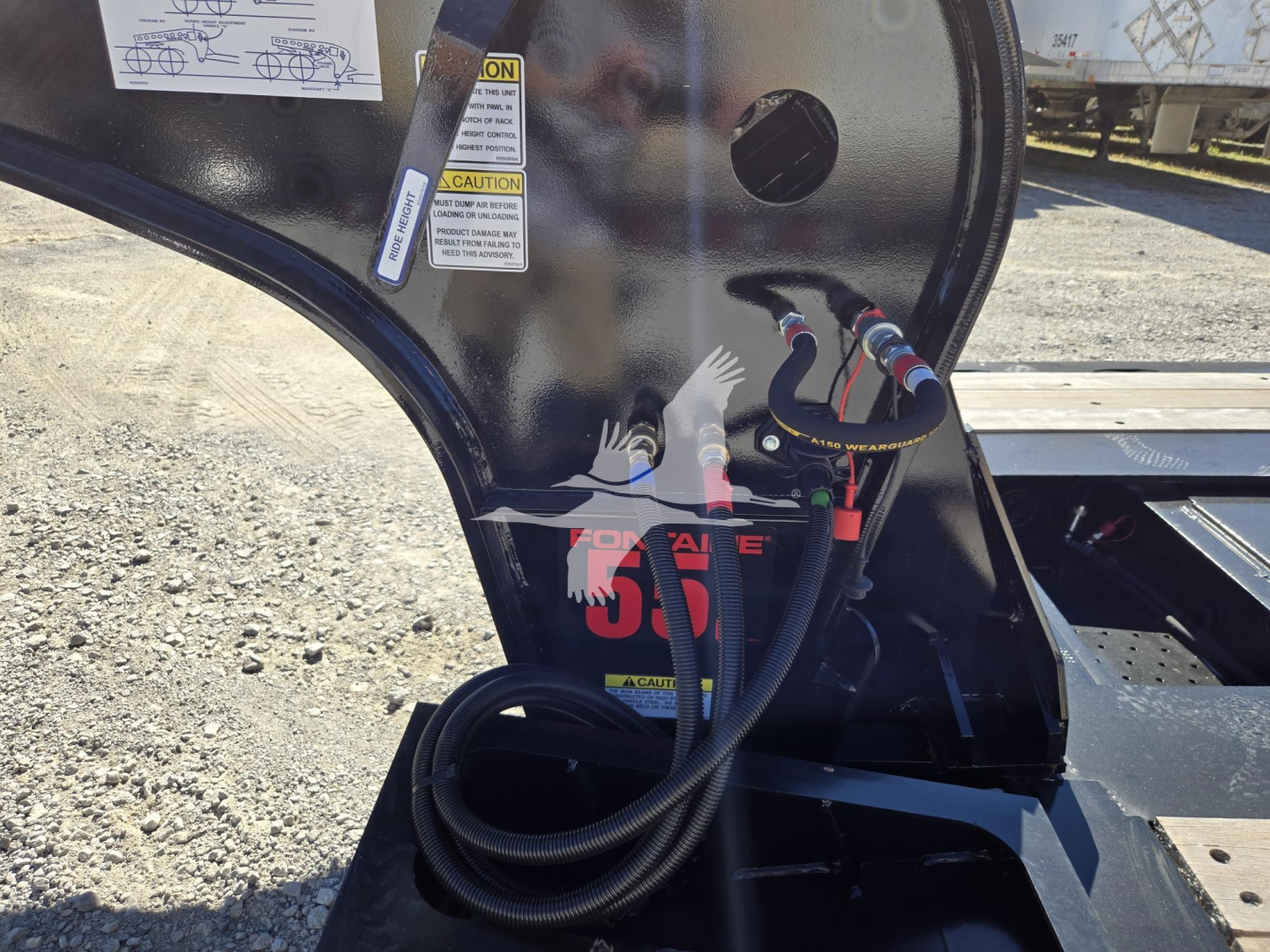 Fontaine lowboy trailer hitch area with hydraulic hoses and warning labels, view shows cable connections and pivoting frame while new trailer (55 Lcc) sits on gravel.