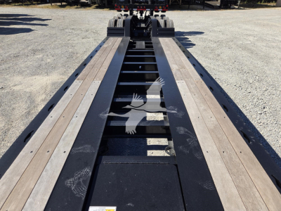 Lowboy trailer deck with long steel frame and wooden planks, hydraulic flip mechanism visible at far end, no cab in view.