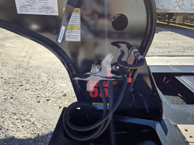 Fontaine lowboy trailer hitch area with hydraulic hoses and warning labels, view shows cable connections and pivoting frame while new trailer (55 Lcc) sits on gravel.