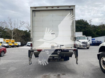 Rear view of a box truck showing a white roll-up cargo door, taillights, and support legs. Visible text includes a small plate or sticker on the lower left area and a rear bumper.