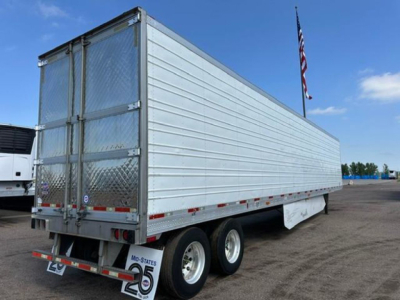 Utility trailer in side view with metallic ribbed exterior and dual rear doors; has triple axles with tires; American flag in background; stock number and VIN not visible on trailer.