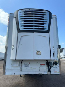 Refrigerated trailer rear view with cooling unit and doors; visible louvered condenser grille, white exterior, various safety stickers near the bumper.