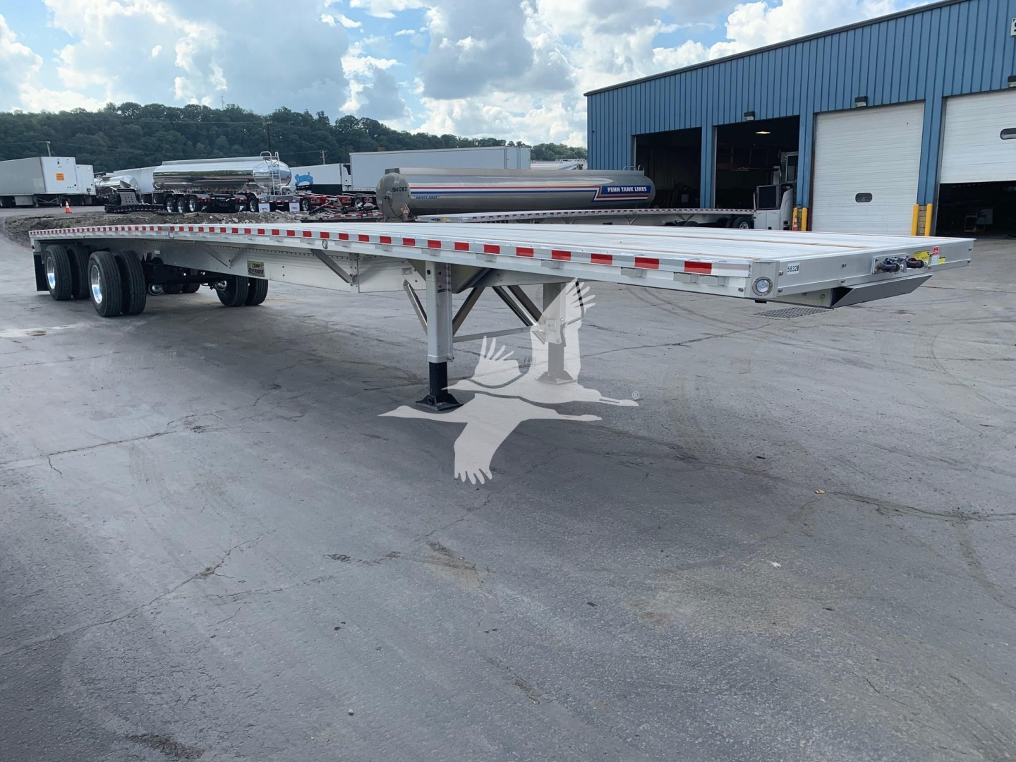 Flatbed trailer with dual rear axles and adjustable landing gear; aluminum deck with red and white reflective tape along sides; blue industrial building in background.