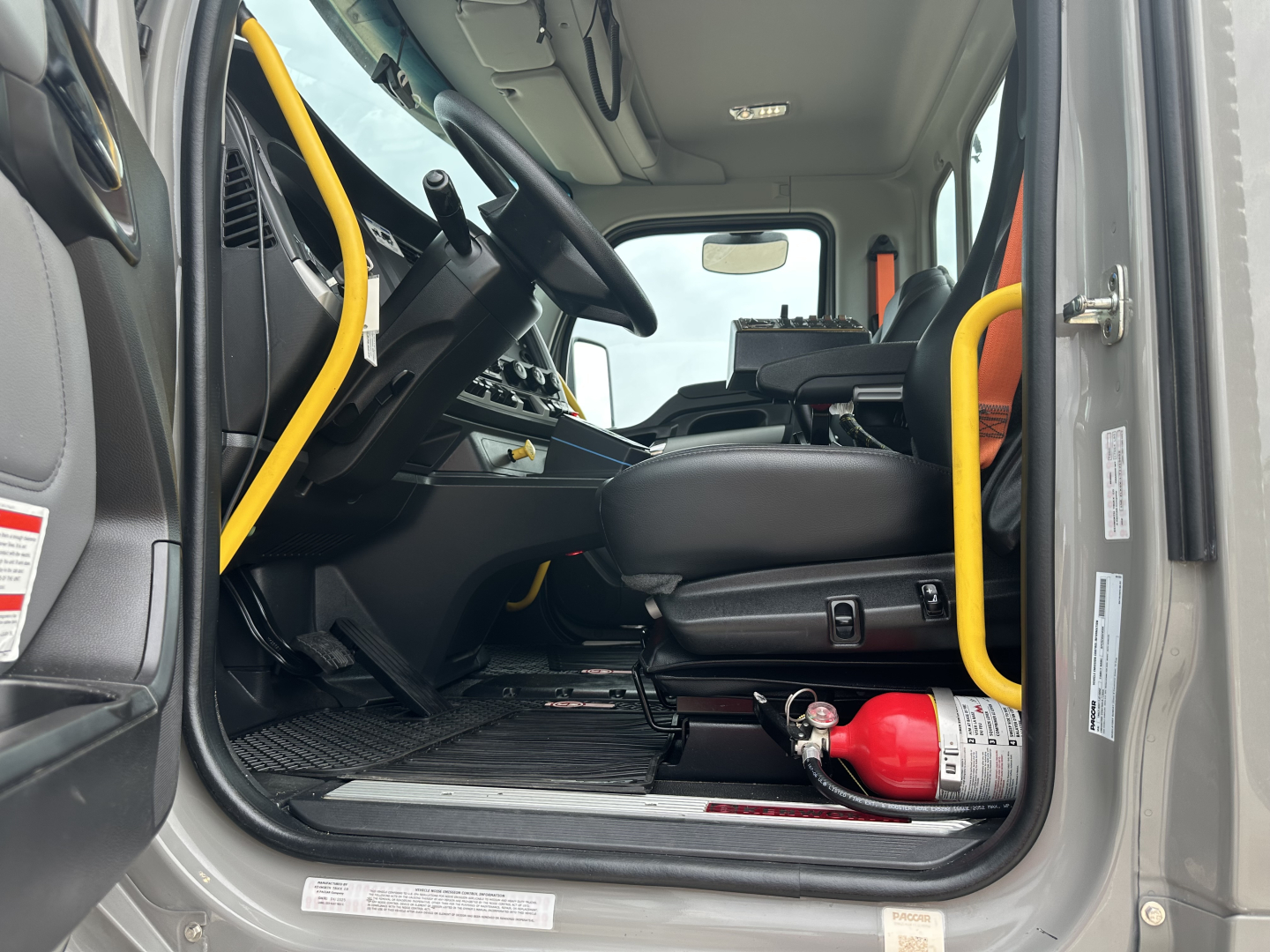 Kenworth T880 mixer truck cockpit interior with steering wheel, dashboard controls, yellow grab handles, black seat, and a red fire extinguisher near the door.