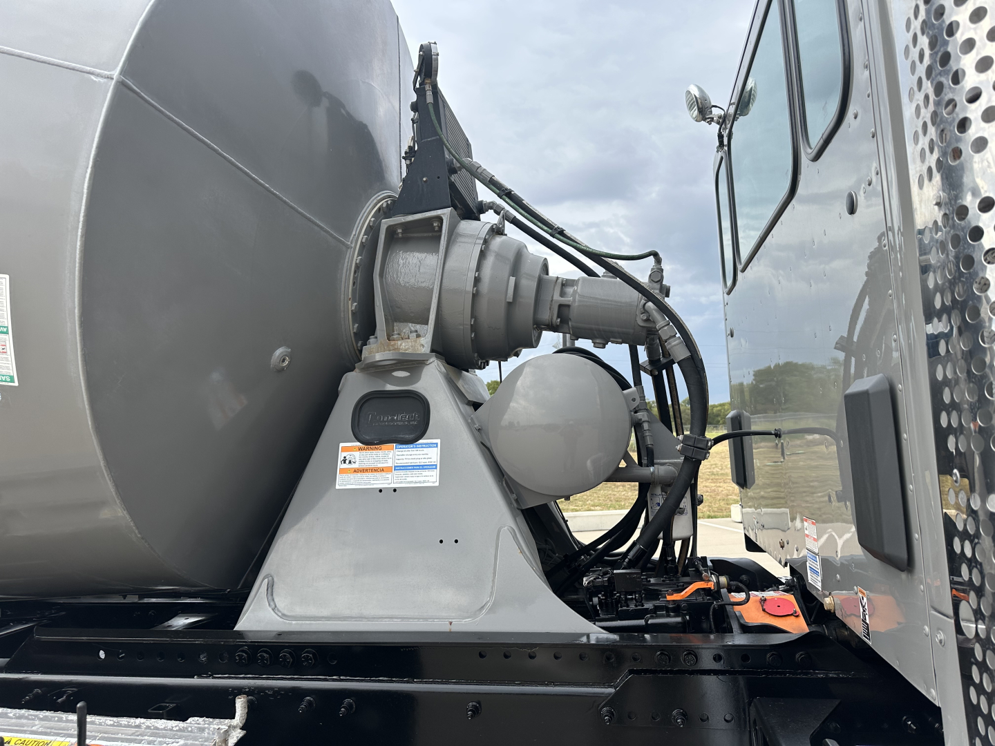 Kenworth mixer truck cab area showing hydraulic hose connections and mounting bracket for mixer drum. Visible labels on brackets, no legible small print.