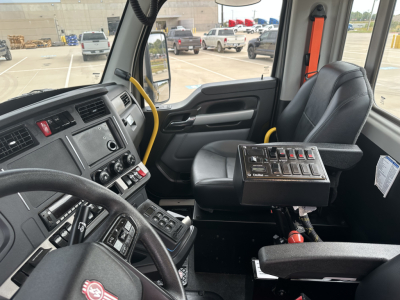 Kenworth T880 cab interior with steering wheel, dashboard, and control panel; yellow handrails; driver seat; window shows trucks in lot.