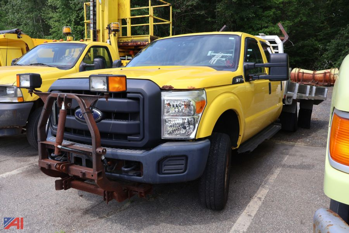 Front view of a yellow 2015 Ford F-350 Super Duty stake truck with a large black grille guard, orange cab marker light, and a rusted front bumper attachment; another yellow truck and equipment trailer visible in background.