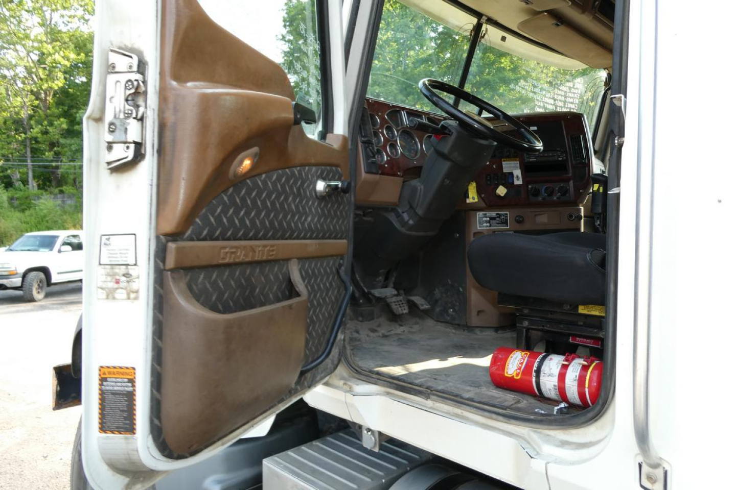 Truck cab interior with open door showing steering wheel and dashboard; brown entry panel, black seat, red extinguisher on floor, metal steps, and door panel with textured pattern.
