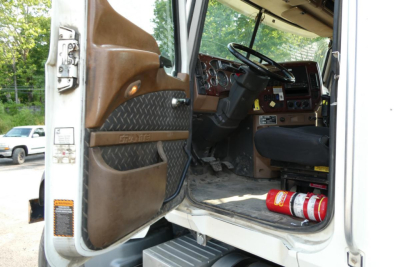 Truck cab interior with open door showing steering wheel and dashboard; brown entry panel, black seat, red extinguisher on floor, metal steps, and door panel with textured pattern.