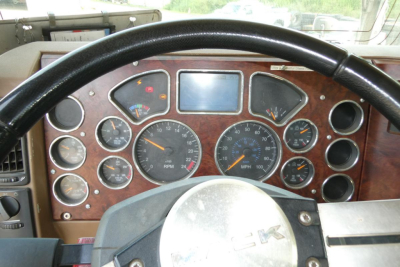 Dashboard view of a Mack GU713 truck showing multiple gauges, speedometer, tachometer, and a wood-grain panel with round dials and a steering wheel.