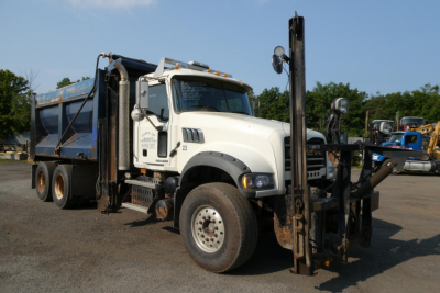 Dump truck front-side view showing Mack GU713 with front-mounted forklift spreader attachment and dump bed. Vehicle white cab, blue dump body, gravel lot.