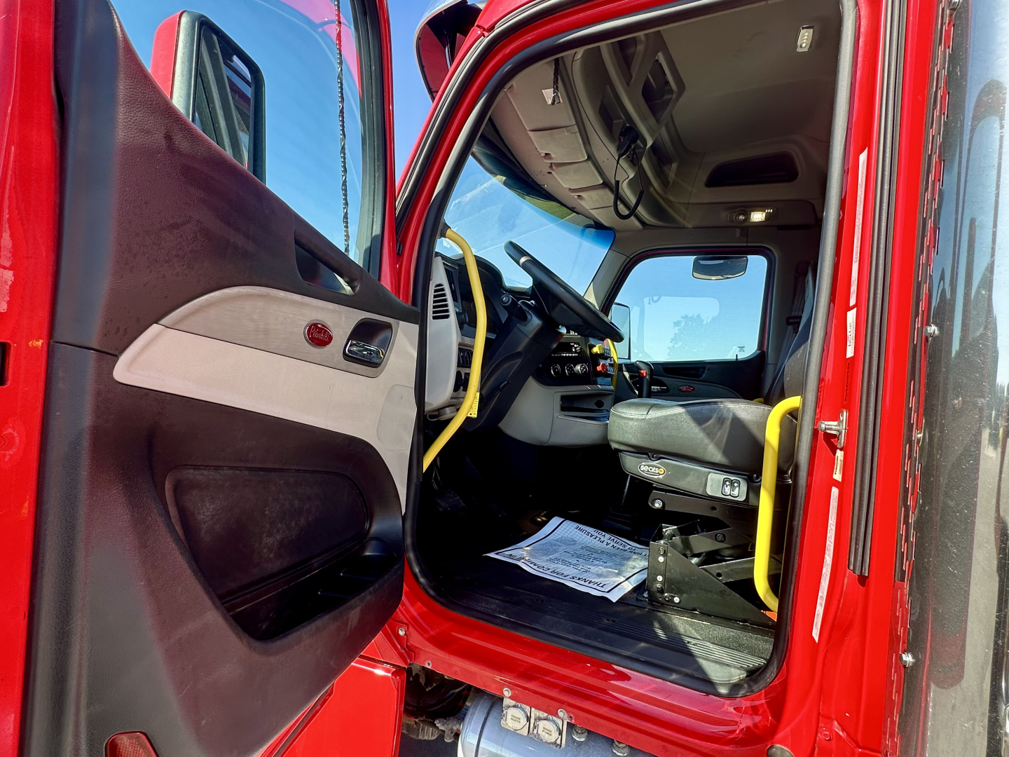 Peterbilt truck cab interior showing steering wheel, dashboard, and entry door with yellow grab handle.
