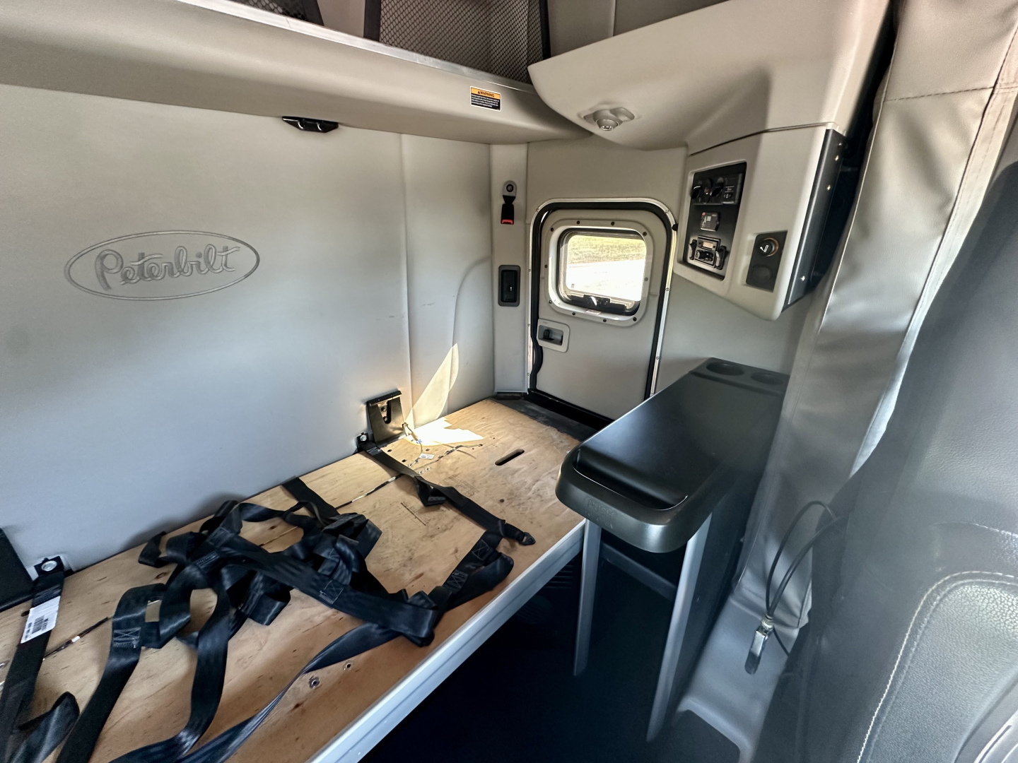 Peterbilt truck cab interior showing sleeper area with seat and window, Peterbilt emblem on wall, seating belts, and side door.