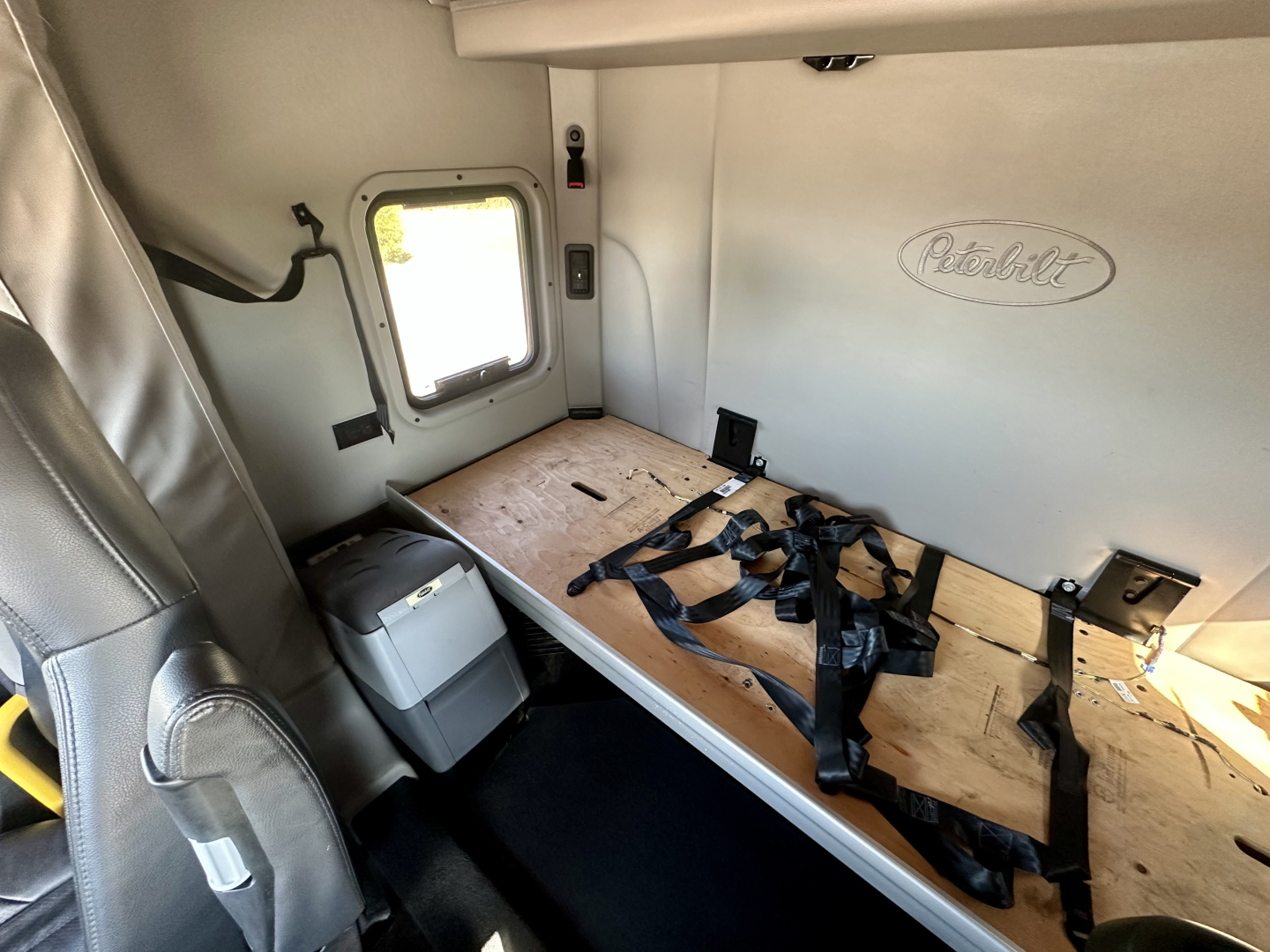 Peterbilt 567 cab interior showing sleeper bunk with wood platform and seat belts, window, and Peterbilt emblem on wall.