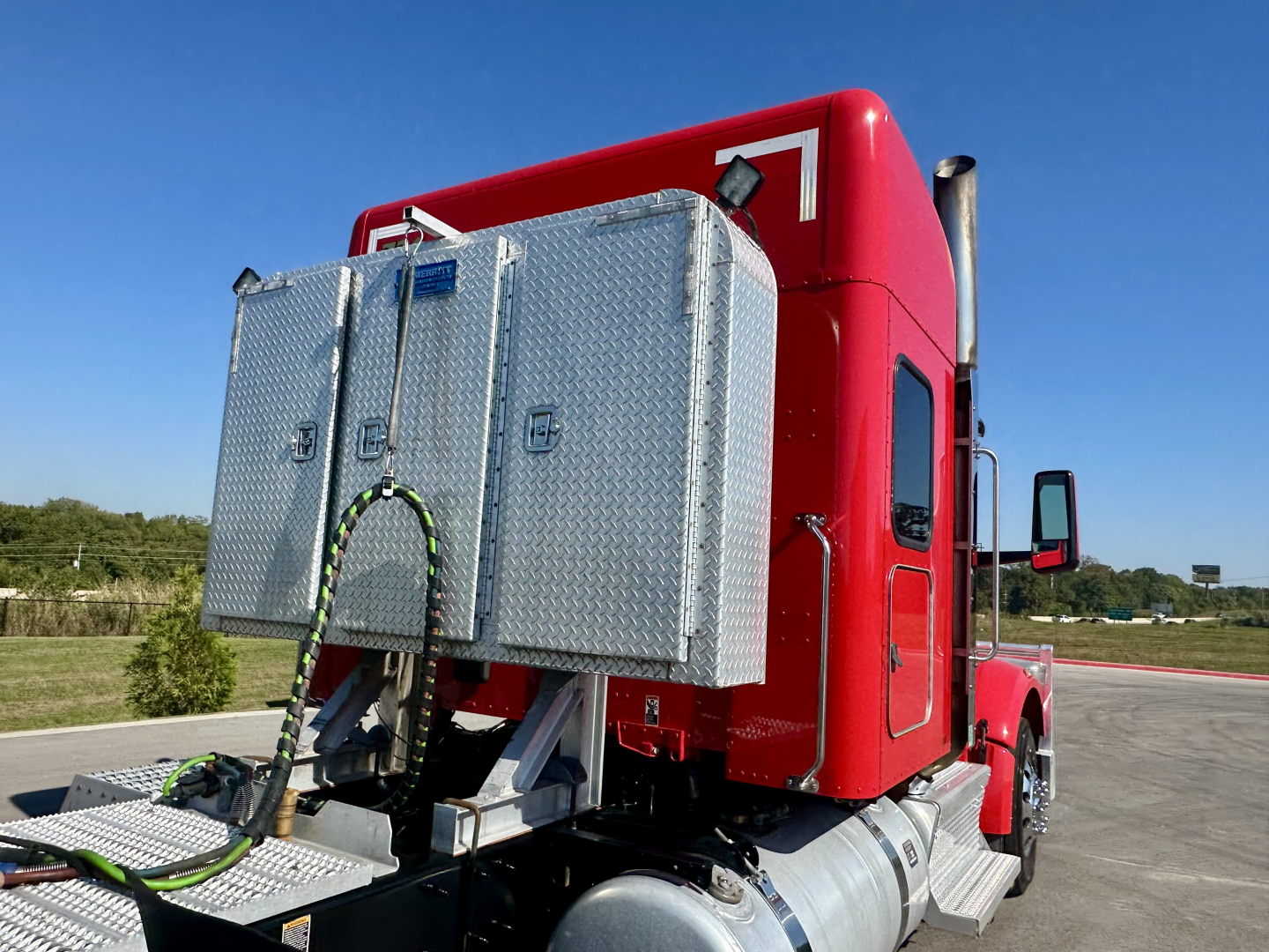 Peterbilt 567 sleeper cab rear view showing aluminum tool boxes mounted behind cab, red exterior, chrome exhaust, and side steps.
