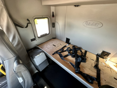 Peterbilt 567 cab interior showing sleeper bunk with wood platform and seat belts, window, and Peterbilt emblem on wall.