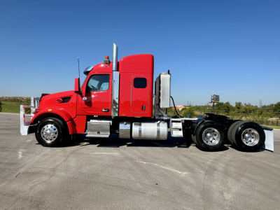 Peterbilt 567 conventional sleeper truck side view, red cab, dual rear axles, chrome fuel tanks and exhaust stack, daytime outdoor lot.