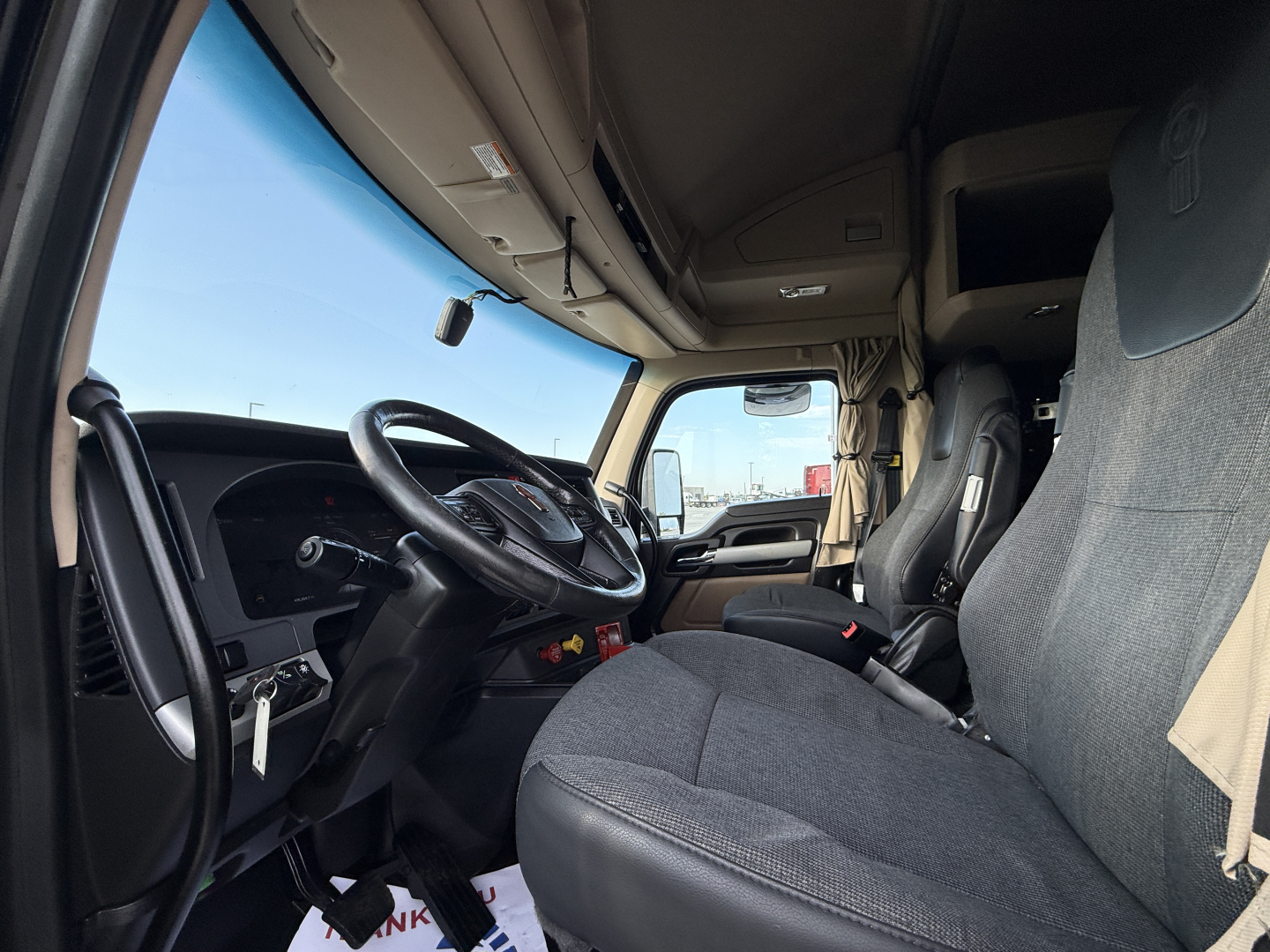 Cab interior of a 2023 Kenworth T680 truck showing steering wheel, dashboard, and driver seat with fabric upholstery and overhead compartments.