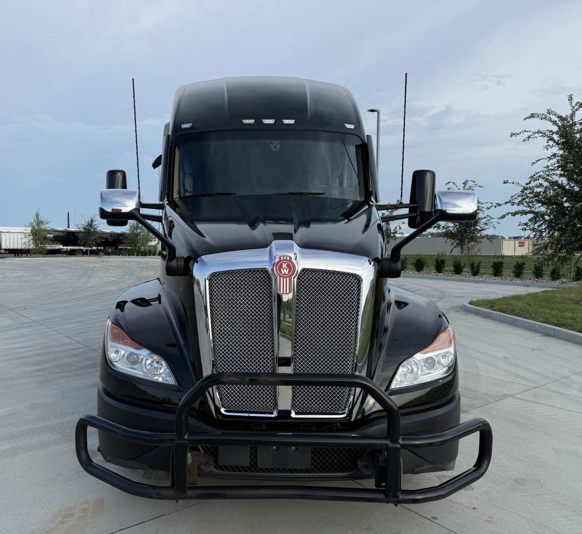 Kenworth T680 truck front view showing large chrome grille, Kenworth emblem, black bull bar, side mirrors, and cab roof fairing.