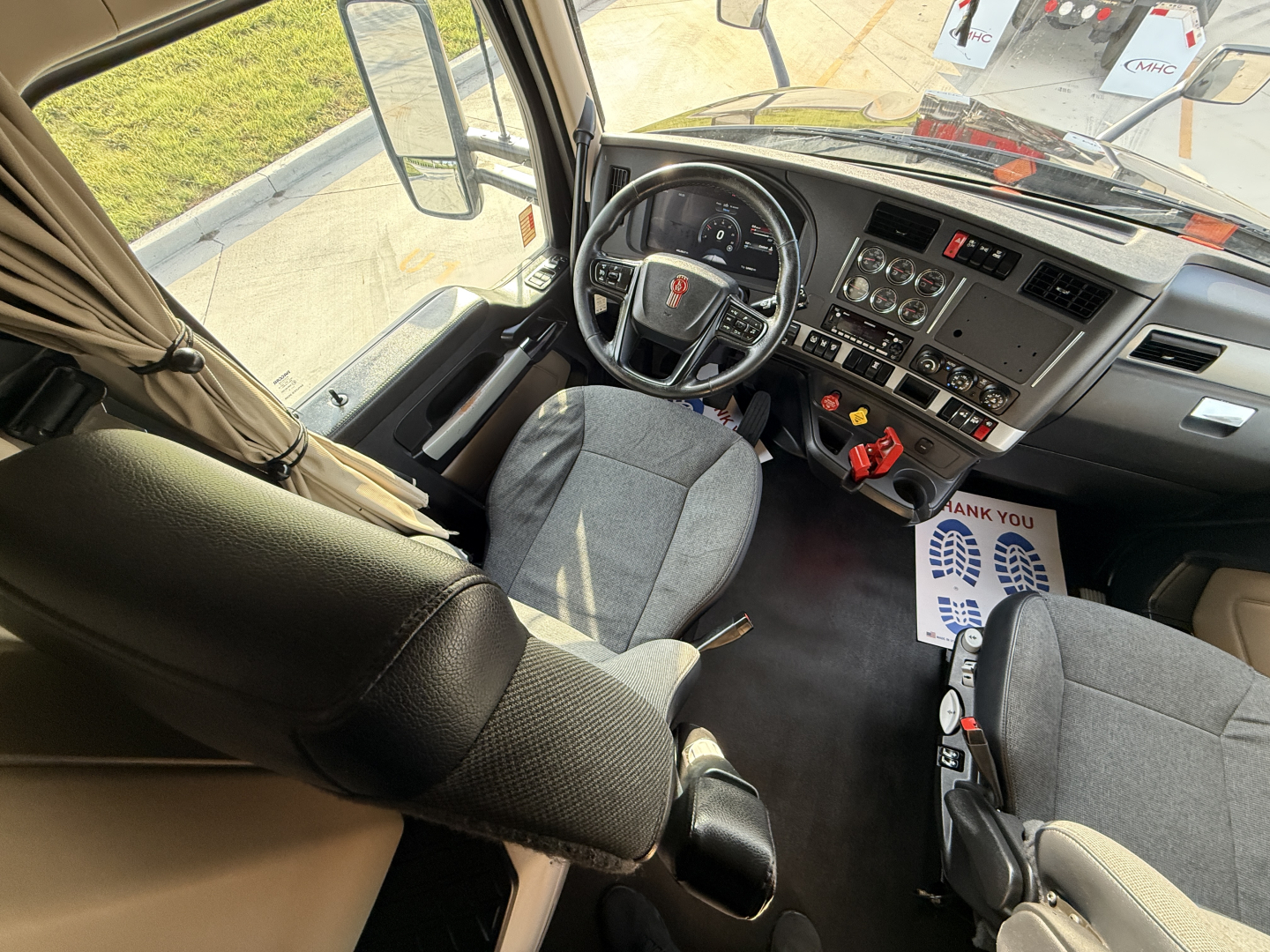 Kenworth T680 truck interior showing steering wheel, dashboard with gauges, center console, and driver seat; light gray fabric seats; side window and door panels visible.