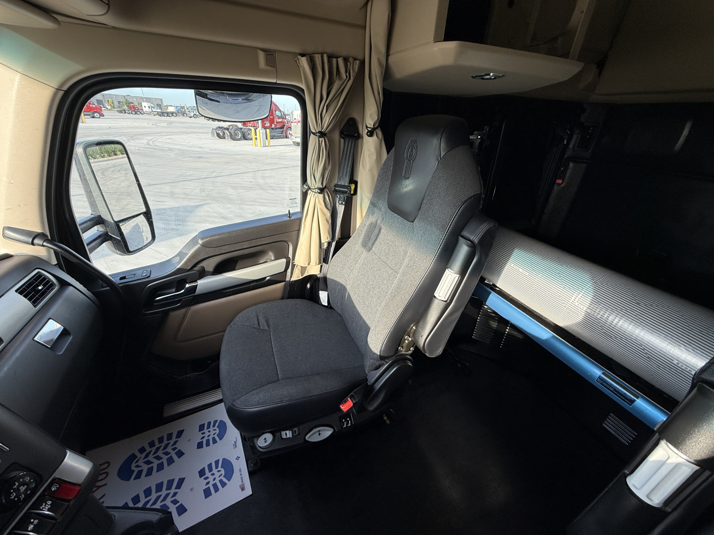 Cab interior of a 2023 Kenworth T680 showing driver seat with steering wheel, side window, dashboard accents and a blue safety prop in the sleeper area.