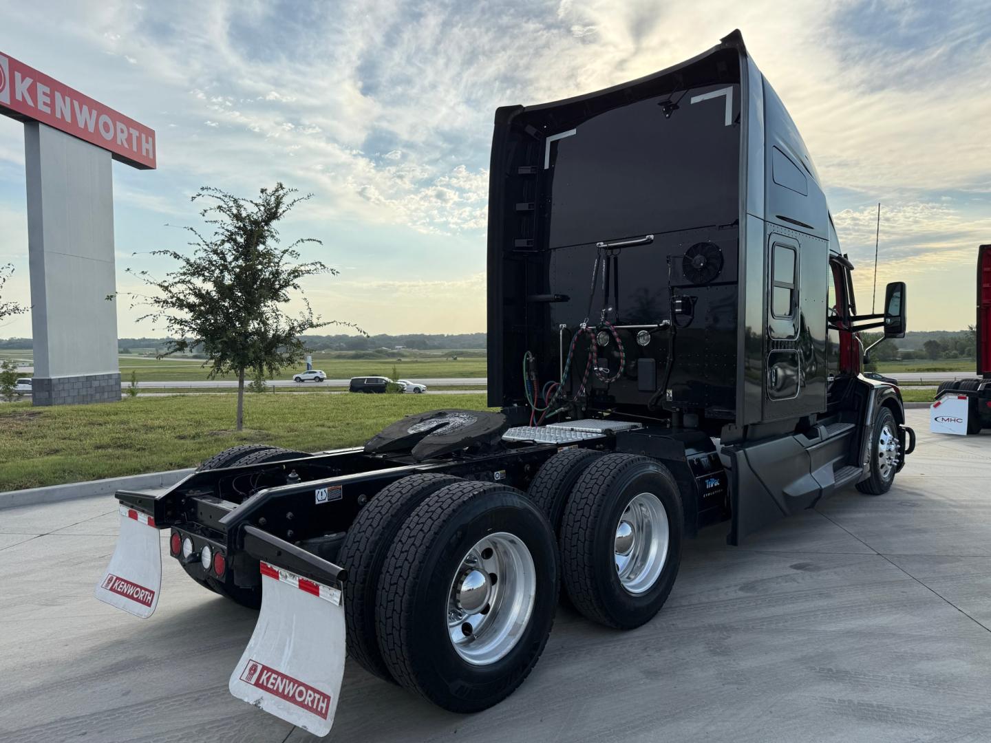 Kenworth T680 tractor unit in dark color, rear view showing dual rear axles and fifth-wheel hitch, with Kenworth dealership sign in background.