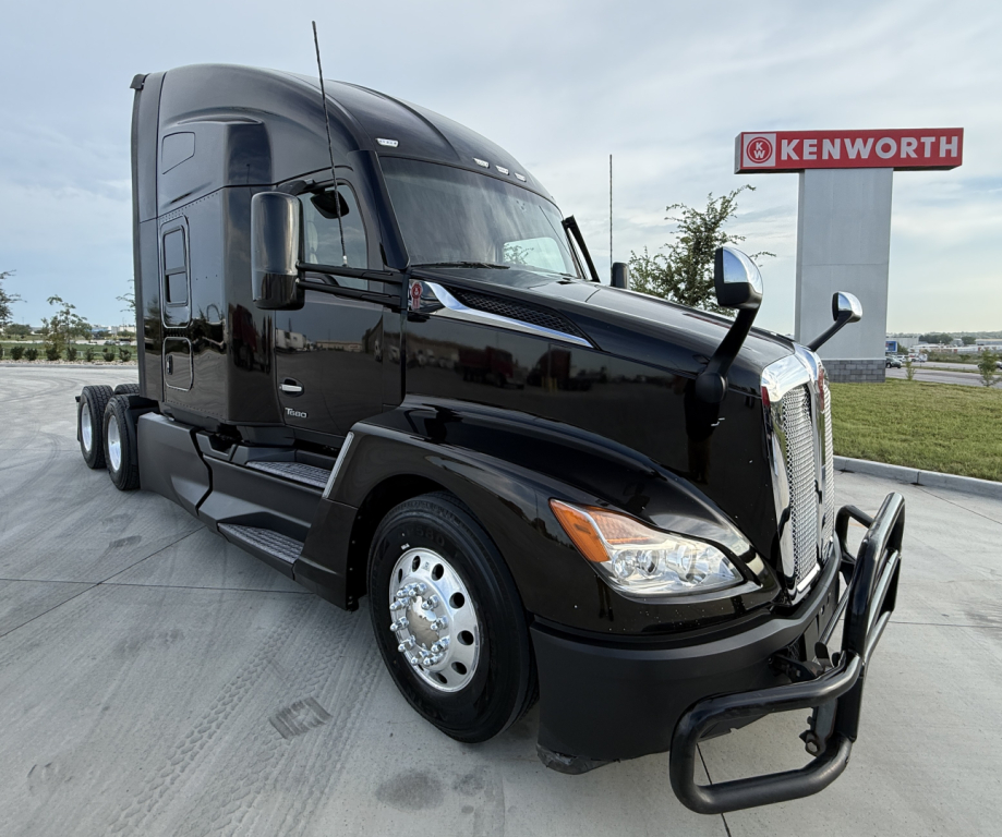 Kenworth T680 semi-truck in black, front-right view showing sleeper cab, chrome wheels, and Kenworth sign in background.