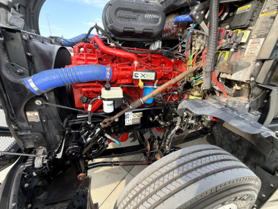 Kenworth T680 engine bay showing red Cummins engine, blue hose, wiring, and various labeled fluid reservoirs.