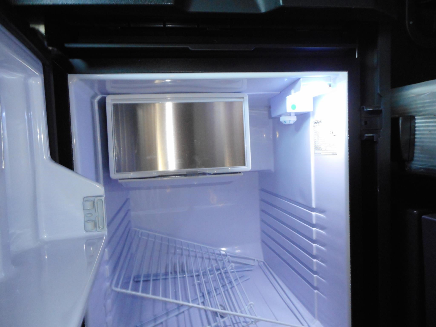 Freightliner Cascadia truck cab interior with empty refrigerator-freezer mounted in small cargo box, illuminated; inside view shows a compact fridge with metal door.