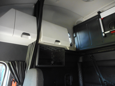 Interior sleeper cabin of a Freightliner Cascadia showing upper storage compartments and a mounted black case with white latches, overhead shelves, and window curtain.