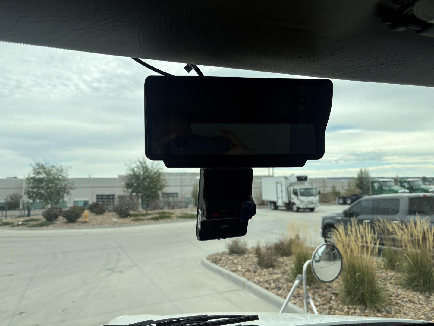Rearview mirror and dash-mounted device in Kenworth T270 cab with windshield view of lot and vehicles.