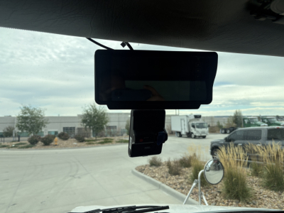 Rearview mirror and dash-mounted device in Kenworth T270 cab with windshield view of lot and vehicles.