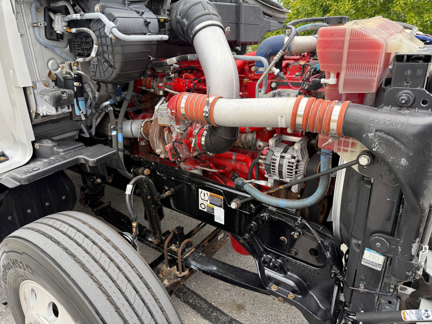 Kenworth T680 engine bay with red engine block, intake piping, coolant reservoir, and various hoses visible; front suspension and frame components in view.