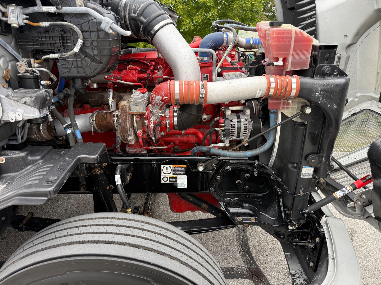 Kenworth T680 engine bay showing red engine block, hoses, coolant reservoir, and air intake pipes in front of the left wheel well.