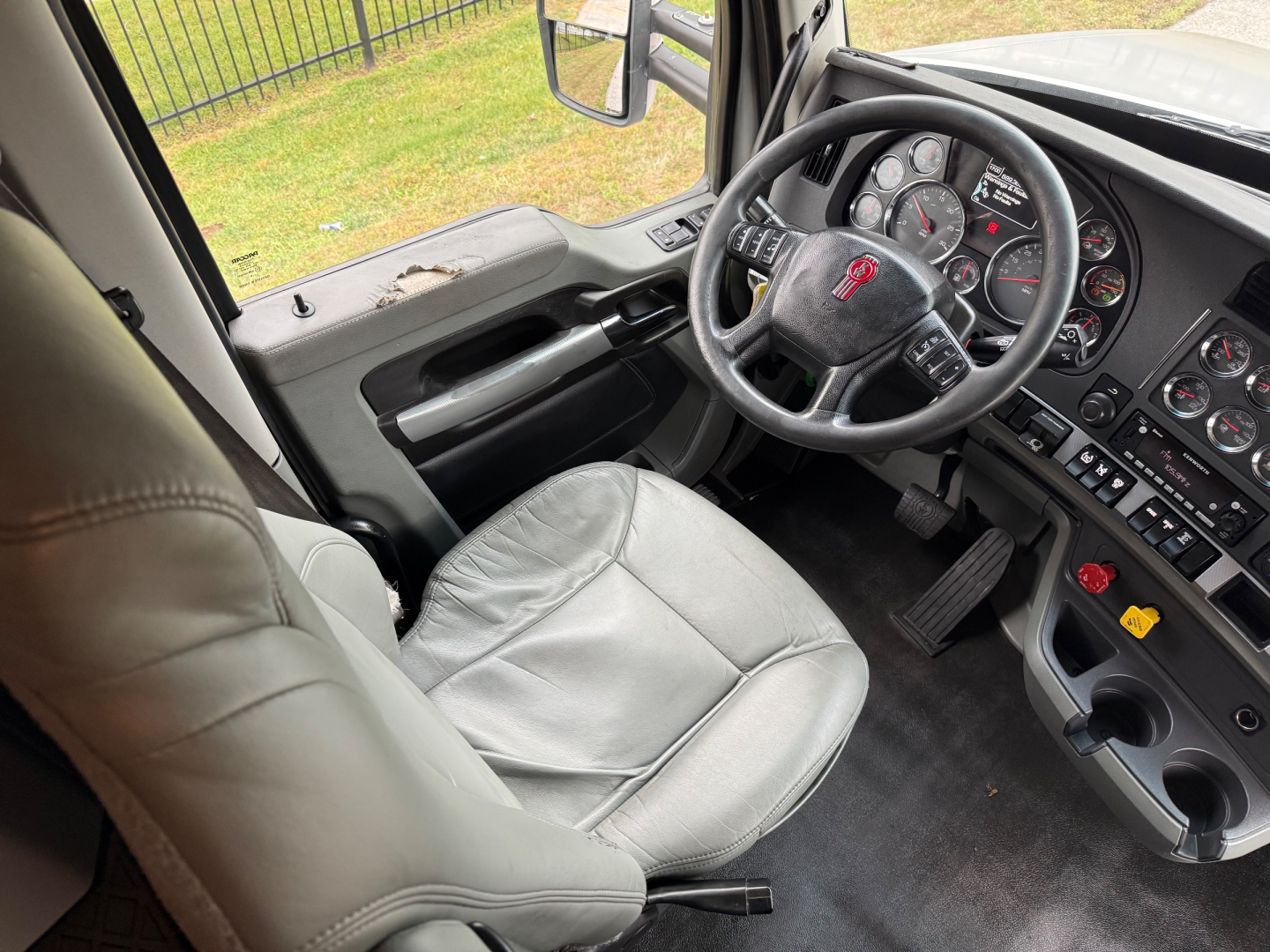 Truck interior showing steering wheel with logo, dashboard gauges, center console, and gray driver seat in a Kenworth T680 cab.