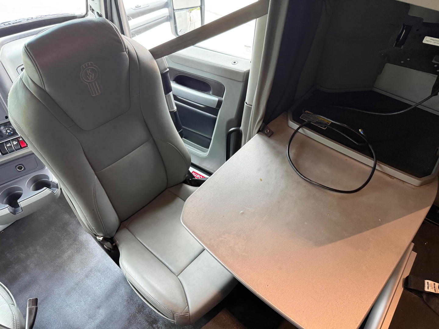 Cab interior of a 2020 Kenworth T680 with a gray captain seat and a fold-out wooden table, dash controls visible, no text legible aside from a chair logo.