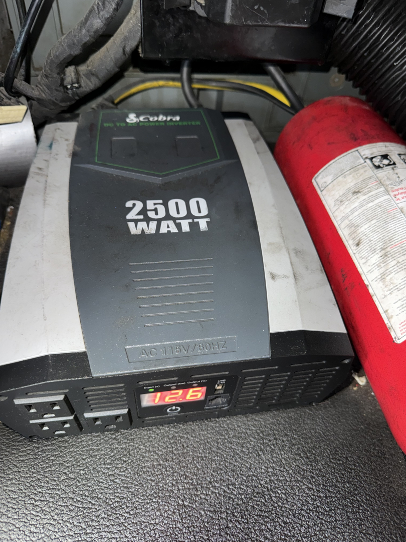 In-vehicle view of a 2500-watt Coburn? inverter labeled 2500 WATT and 12V; visible control panel with gauge; red fire extinguisher beside; wiring and components in the engine bay area.
