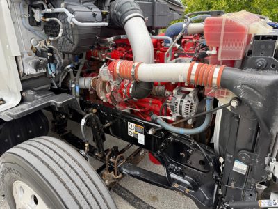 Kenworth T680 engine bay with red engine block, intake piping, coolant reservoir, and various hoses visible; front suspension and frame components in view.