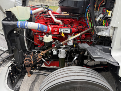 Kenworth T680 engine bay with red ISX engine, various hoses, coolant reservoir, air lines, and wiring visible.
